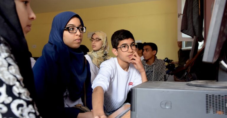 Two teenagers, a girl with a headscarf and a boy, both wearing glasses, look at the screen of a desktop computer in a classroom.