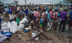 A large group of Ugandan citizens in a displaced person’s camp. Many are standing around talking or sitting on or near bags and buckets.]