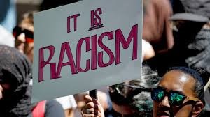 A person with dark skin wearing sun glasses at a protest carrying a white sign with pink letters that reads IT IS RACISM.
