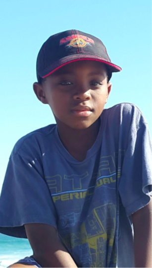 A young, Black South African boy wearing a blue t-shirt and a black cap, looking straight at the camera.