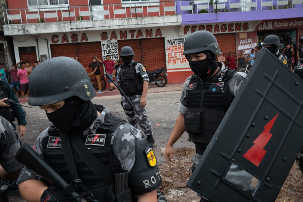 Brazilian police in riot gear with machine guns, helmets, and shields on one side of a town’s street with civilians on the other side of the street.]