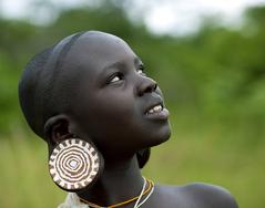 A Kenyan girl with dark skin, a shaved head and a stretched earlobe with a large patterned disc in her earlobe. The disk has 5 concentric, uneven brown circles on a bone colored background and the edges have small brown rectangles all around. She is looking upward toward the sky and smiling.