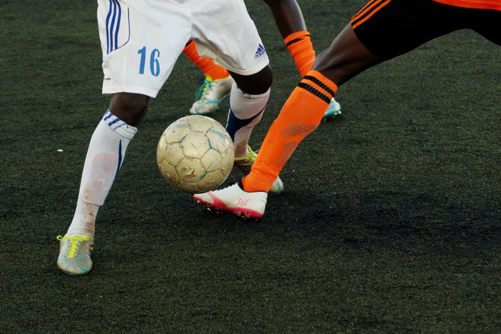 The legs and feet of three football/soccer players while playing the game. One player has white shorts, white socks and white shoes. The other two have black shorts, orange socks, and white shoes. One of the players with black shorts is trying to kick the ball through the legs of the opposing player. All players are Black with dark skin.
