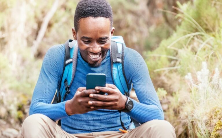 A youth wearing a backpack smiles into a mobile phone while sitting outdoors