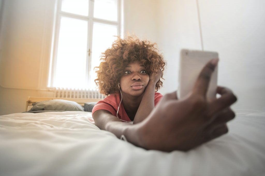 A Black young woman with mid-length wavy light red hair leaning prone on a bed, looking at her phone with an earbud wire coming down from her ear.