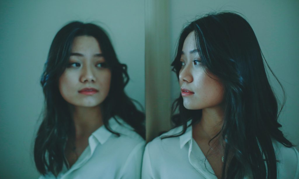 An Asian woman with long black hair and red lipstick wearing a white collared button down shirt and her reflection as she looks in a mirror.