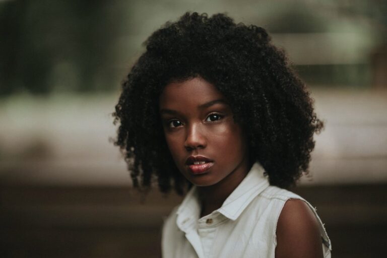 A Black young woman with dark skin and long black curly hair wearing a white sleeveless button-down collared shirt looking directly at the camera.