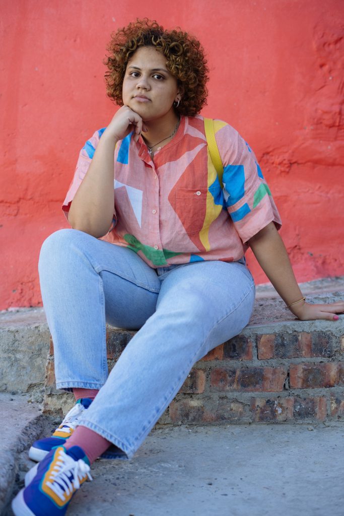 A woman with light brown skin, curly hair and hoop earrings. She is wearing a pink, collared button-down shirt with large geometric shapes in various primary colors, jeans, pink socks and purple, green and yellow sneakers with white laces. She is sitting on a raised cement sidewalk built up with red bricks in front of a wall painted reddish-pink with her right knee bent and her elbow on her knee while her fist props up her chin. She is looking directly at the camera.