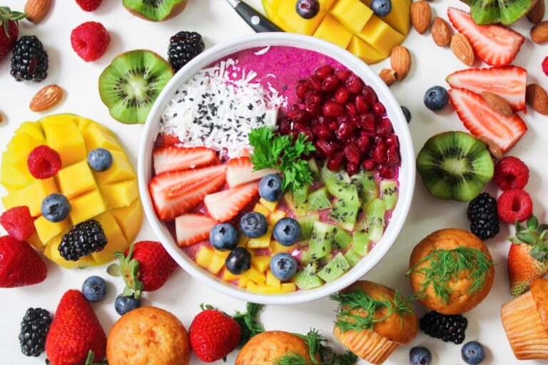 A white bowl of strawberries, blueberries, kiwi, and other fruits is surrounded mangos, nuts, berries, and miniature muffins.