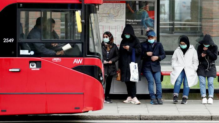 A red bus in front of five people who are standing on a sidewalk wearing coats, head coverings and face-masks.