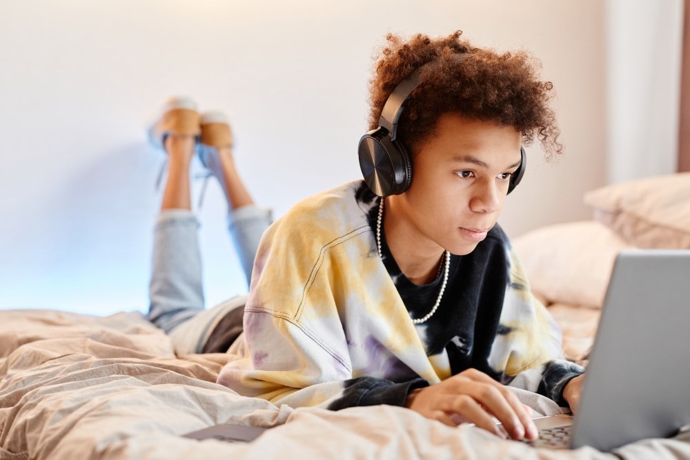 A youth with headphones lies on a bed while using a laptop computer