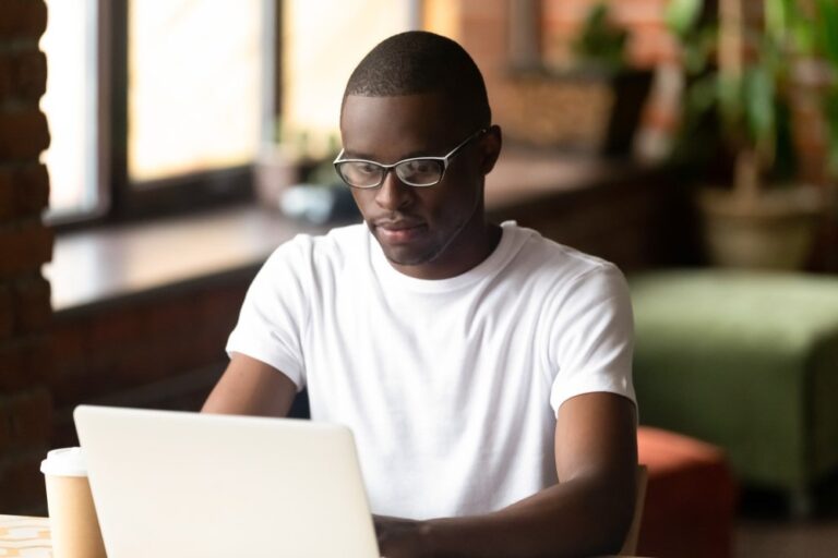 A dark-skinned youth with glasses and a t-shirt works on a laptop computer