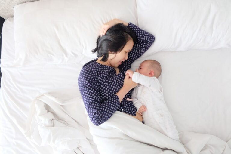 This image shows a young Asian mother in an indigo pajama shirt lying with her infant, who is dressed in white pajamas, in a bed with a white sheet. The mother is awake while smiling at her baby, who is holding her thumb.
