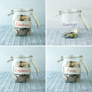 Four open glass jars labeled "donations," "savings," "emergency," and "foods" are filled with various amounts of coins. Donations, emergency, and foods jars are almost full with coins while savings has only about 1/5 of the jar filled. The jars sit on a grey tile-textured table against a white background.