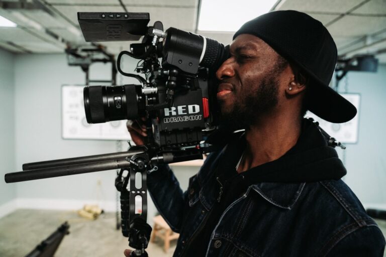 A man with dark skin, a backwards cap, and a jean jacket over a black hoodie is holding a professional camera and looking through it. The background is a studio with multiple cameras and equipment.