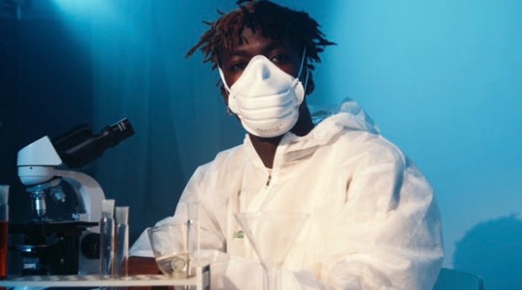 A man with a dark skin tone and short dreadlocks wearing a white lab gown and a white mask next to a microscope and beakers filled with liquid.