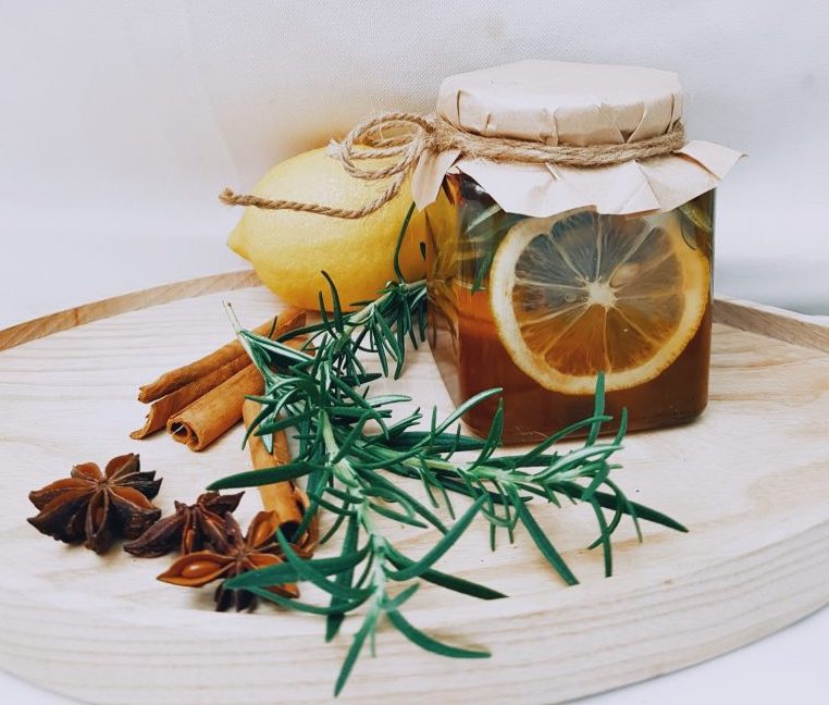 A glass jar with brown liquid, lemon pieces, and rosemary inside with a paper lid on top. it is placed in the middle of a wooden circular tray with yellow lemon, rosemary, and cinnamon beside it.