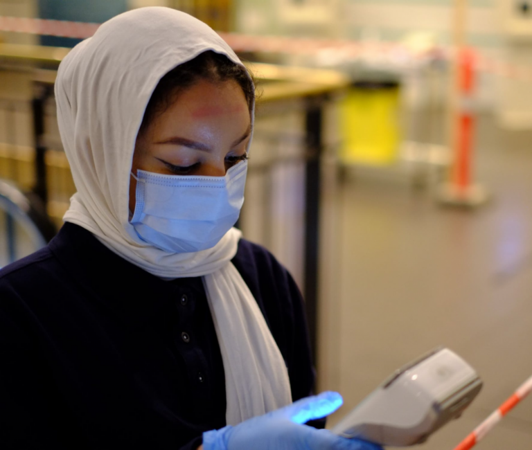 A female wearing a white hijab, a surgical mask, and gloves pressing on a digital thermometer.