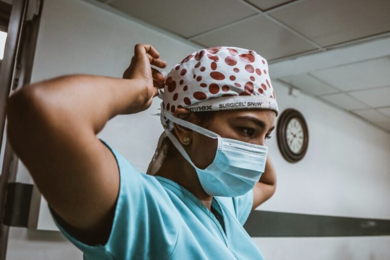 A woman with an olive skin tone wearing light blue scrubs and a white with red dots surgical cap, putting on a blue surgical mask.