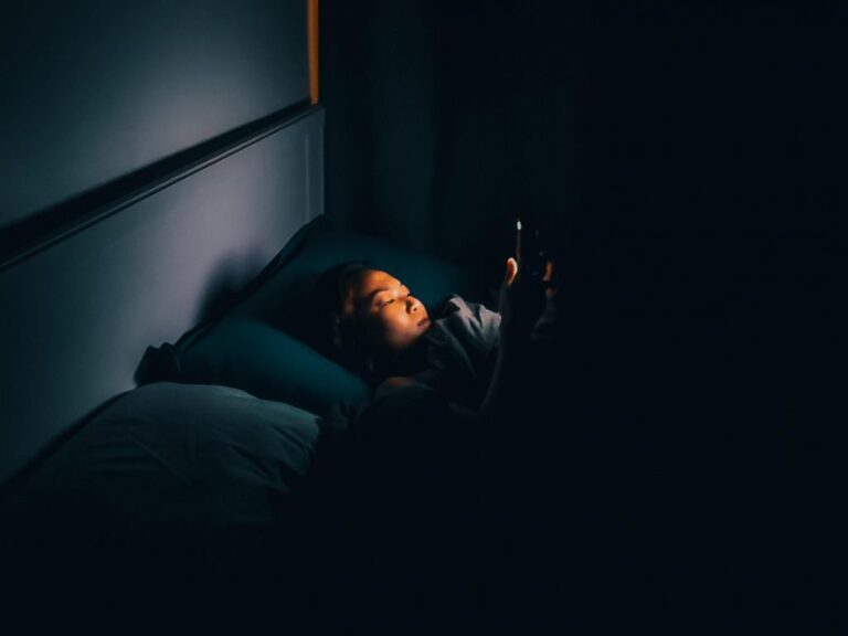 A woman laying down on her bed holding her phone in the dark. She is the only thing illuminated in the dark room